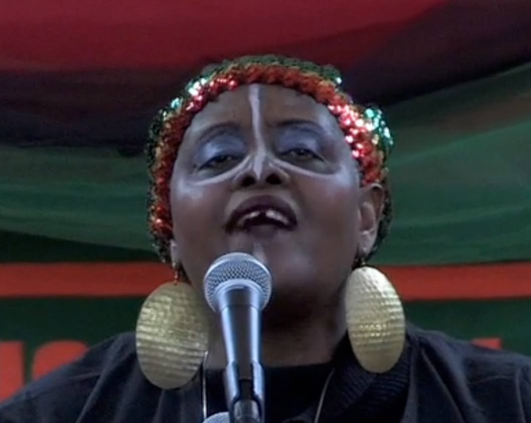 Lola Louis performing as the Griot in The Children's Legacy, in a red, green, and gold sequined headwrap with large gold earrings